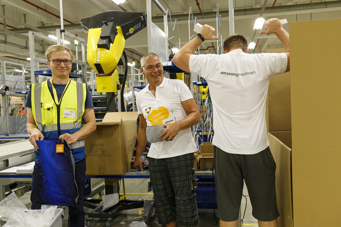 3 Mitarbeiter verpacken Schuluntensilien: Alle 3 lächeln. Ein der Mitarbeiter ist von hinten zu sehen, er deutet mit beiden Daumen auf den T-Shirt-Aufdruck am Rücken "Amazon gemeinsam".