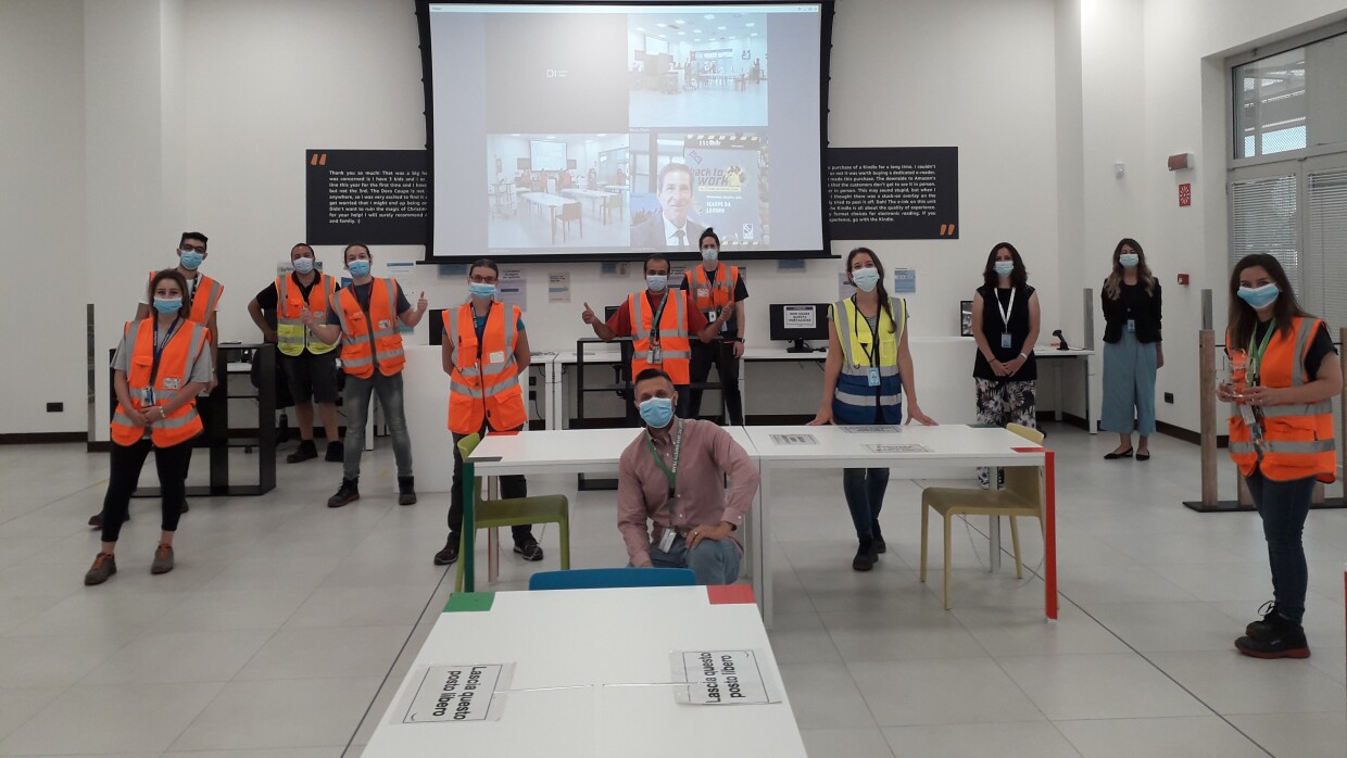 Operatori di magazzino posano in un'area comune del centro di distribuzione Amazon di Vercelli. Indossano la mascherina, sono distanziati.