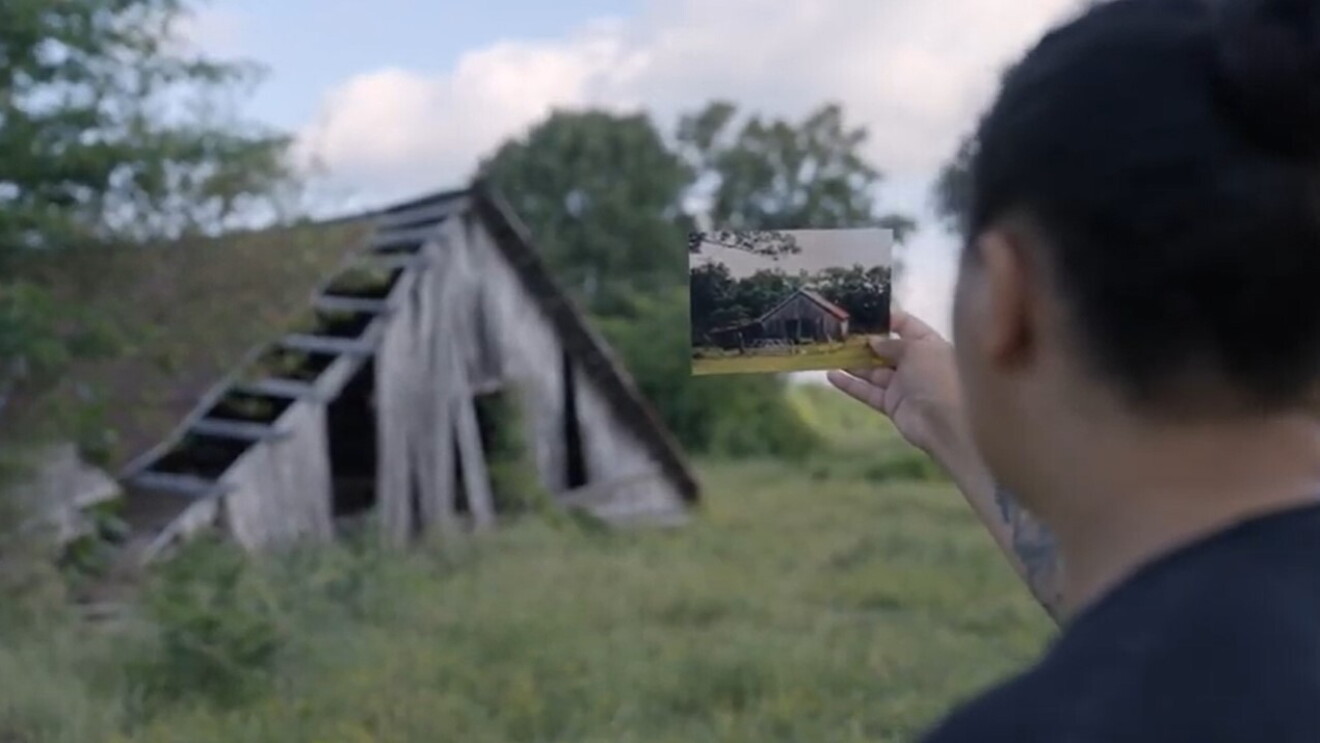 Newton holds up an old photograph of a farm building as she stands in front of the same building, which is now run down.