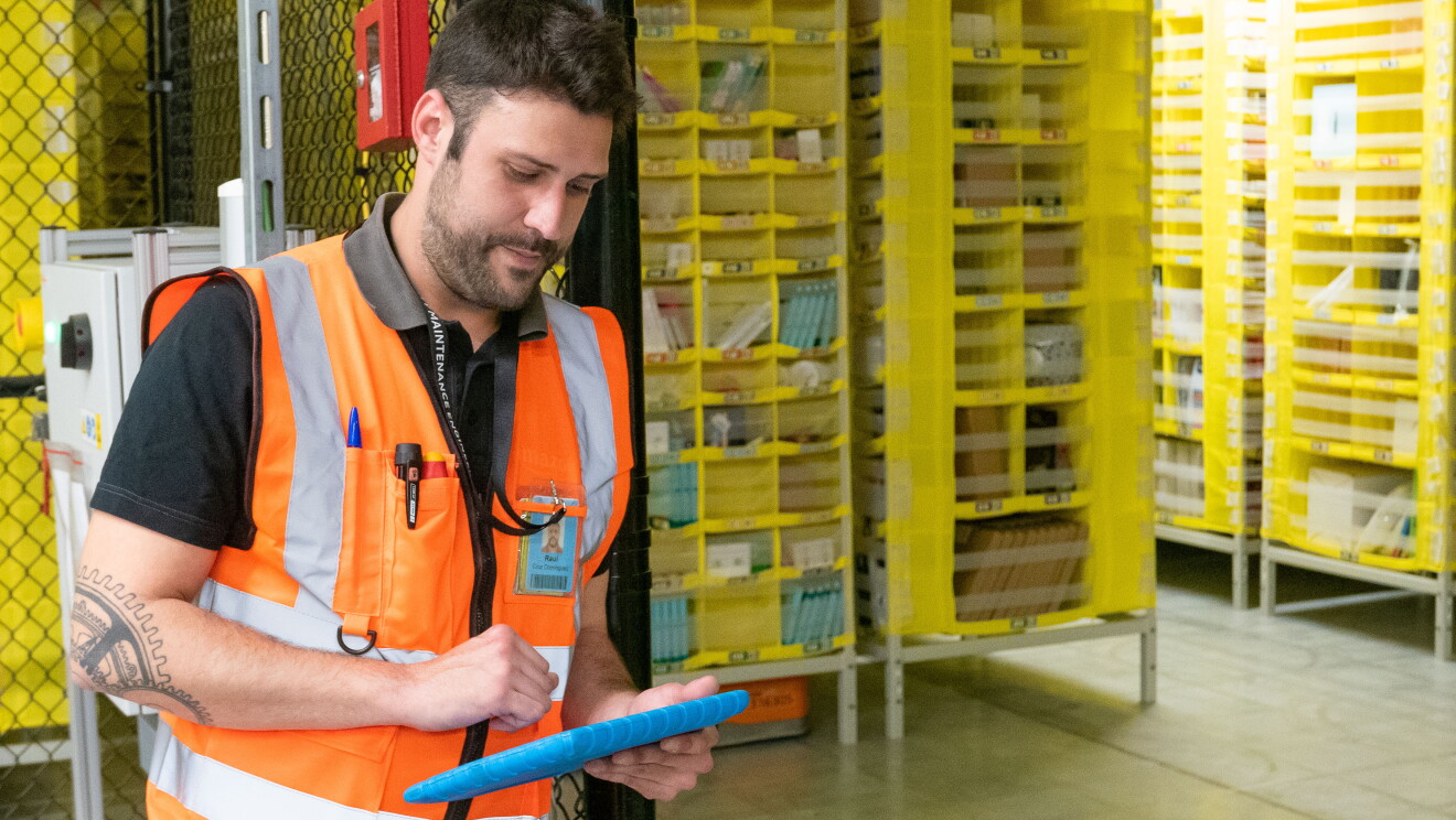 Raúl Cruz, técnico senior de mantenimiento en robótica, con el Kindle controlandos los robots. En primer plano está Raúl, con el pelo negro y barba, vestido con una camiseta negra y el chaleco naraja. En la mano está manipulando un Kindle con una funda azul. De fondo tiene las pods, estanterías que trasladan los robots. 