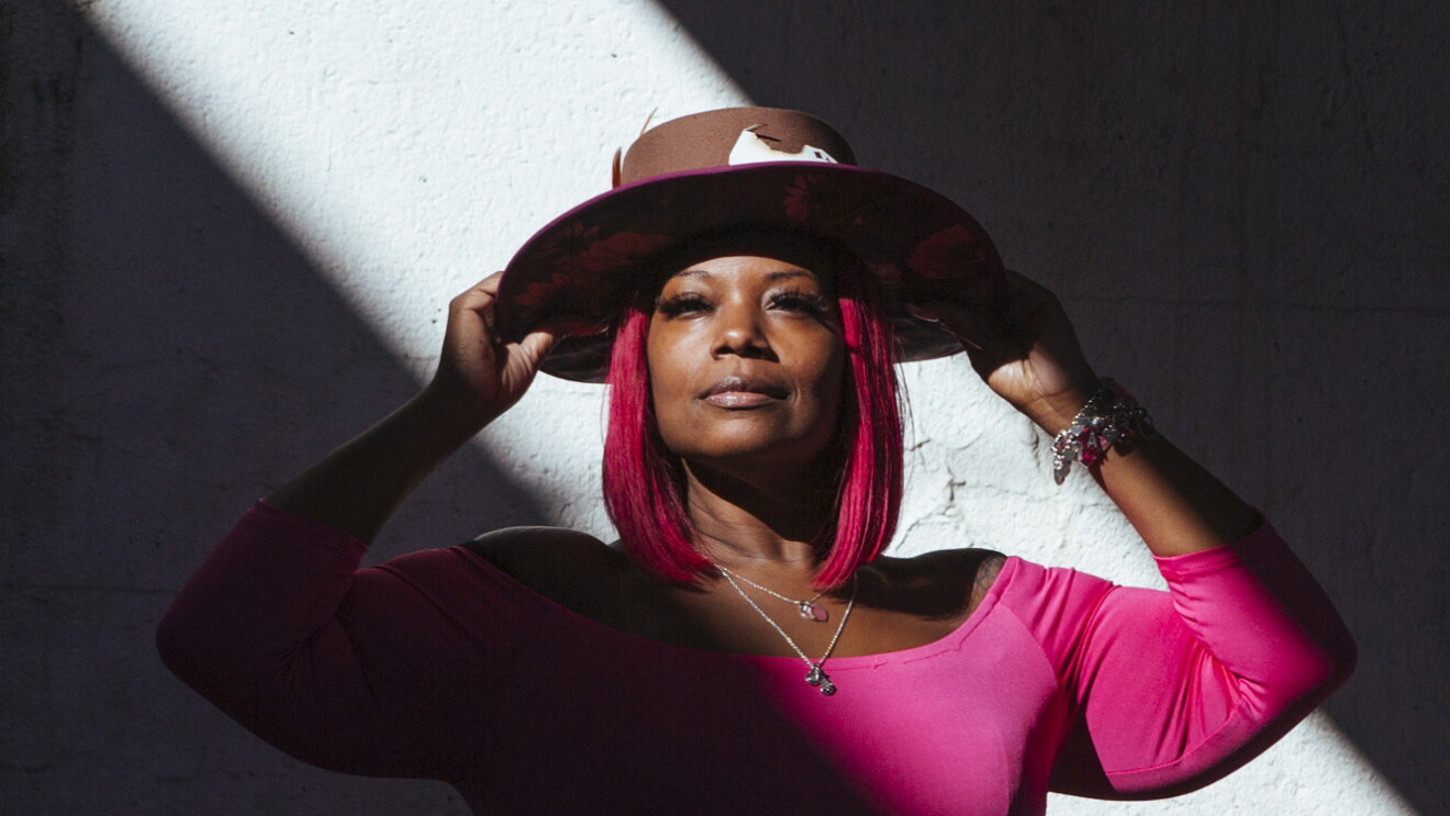 A closeup of Auhzha Wright, with her arms raised and holding her custom-made hat, wears a pink dress and stands between two diagonal shadows.