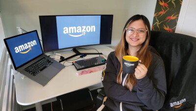InternDay. Una mujer de unos 25 años con el pelo largo de color negro y mechas de color marrón. Lleva una sudadera de Amazon y está agarrando con la mano izquierda una taza de color negro por fuera y amarilla por dentro con el logo de Amazon en color blanco. Está sentada en una silla delante de su mesa y dándole la espalda. En la mesa hay un ordenador portátil conectado a un display port y tiene una pantalla muy grande y un teclado. En las dos pantallas aparece el logo de Amazon. En la parte izquierda hay una barandilla de hierro blanca y en la derecha un cuadro rectangular vertical con un estampado de hojas de color rojo, naranja y verde. 
