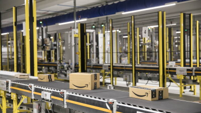Dentro del centro logístico de Amazon en El Prat de Llobregat hay 4 cintas transportadoras que cruzan la foto. Solo en una de ellas, en la que está en primer plano, se ven cuatro cajas de Amazon de diferentes tamaños. En tres de ellas se ve la sonrisa. Las tres cintas transportadoras están separadas por una verjas de color negro y amarillo.  En el techo hay un conducto de color azul. 