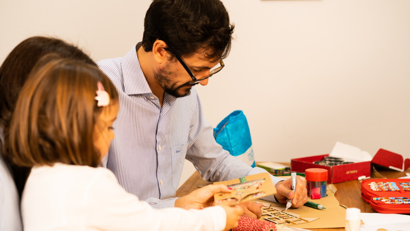 La afición familiar de hacer manualidades aumentó con el confinamiento. En una mesa de salón de madera y llena de objetos para hacer manualidades: hilo, rotuladores, pinceles, pegamento o pegatinas están sentados Fernando, Marta y Lucía. Fernando con una camisa de rayas está pintando una casa de cartón. Marta está tapada por su hija Lucía. Marta le aguanta un cartón mientras Lucía lo manipula. La niña lleva una pinza en el pelo y va con una camisa blanca. 