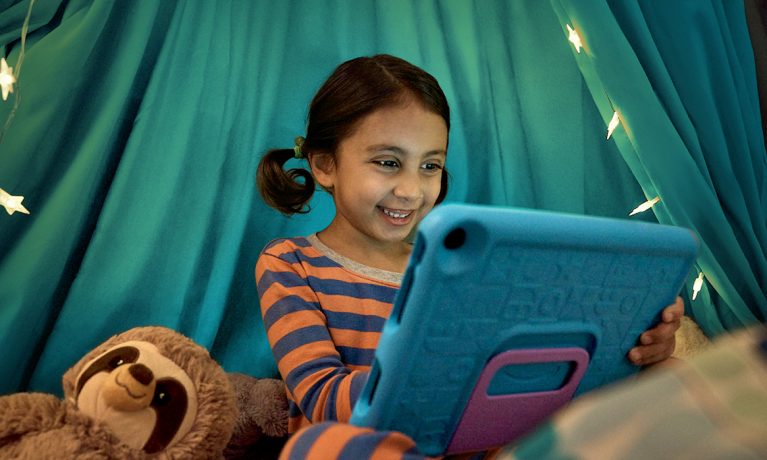 Smiling young girl using kindle device with a sloth stuffed animal on her right