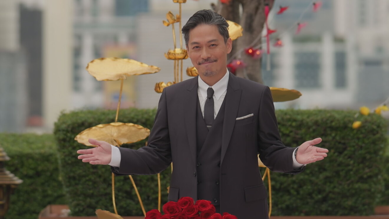 屋外、植木の前で三つ揃いスーツを着た男性が笑顔で両腕を広げたいる。背後に金色のハスのオブジェ。