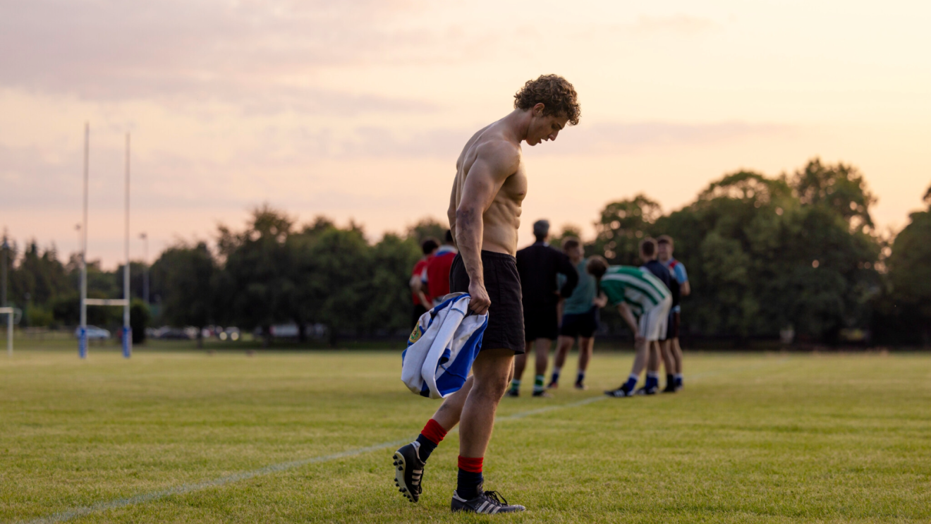 Conor Sánchez (Johnny Kavanagh) on a rugby pitch, shirtless, with his team in the background.