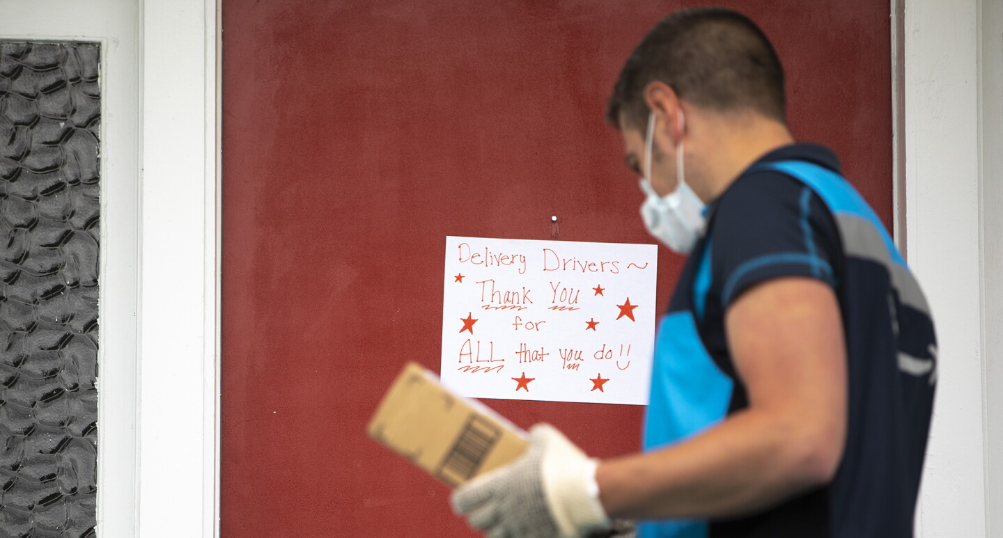 Amazon delivery driver stands in front of a door, to deliver a package. On the door is a sign that says "delivery drivers, thank you for ALL that you do."