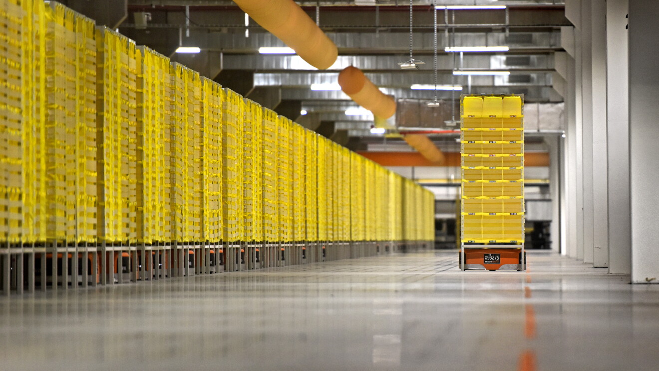 Ein Roboter transportiert ein gelbes Regal durch eine Halle. Links im Bild sind in einer langen Reihe weitere gelbe Regale zu sehen unter den Transportroboter sind.