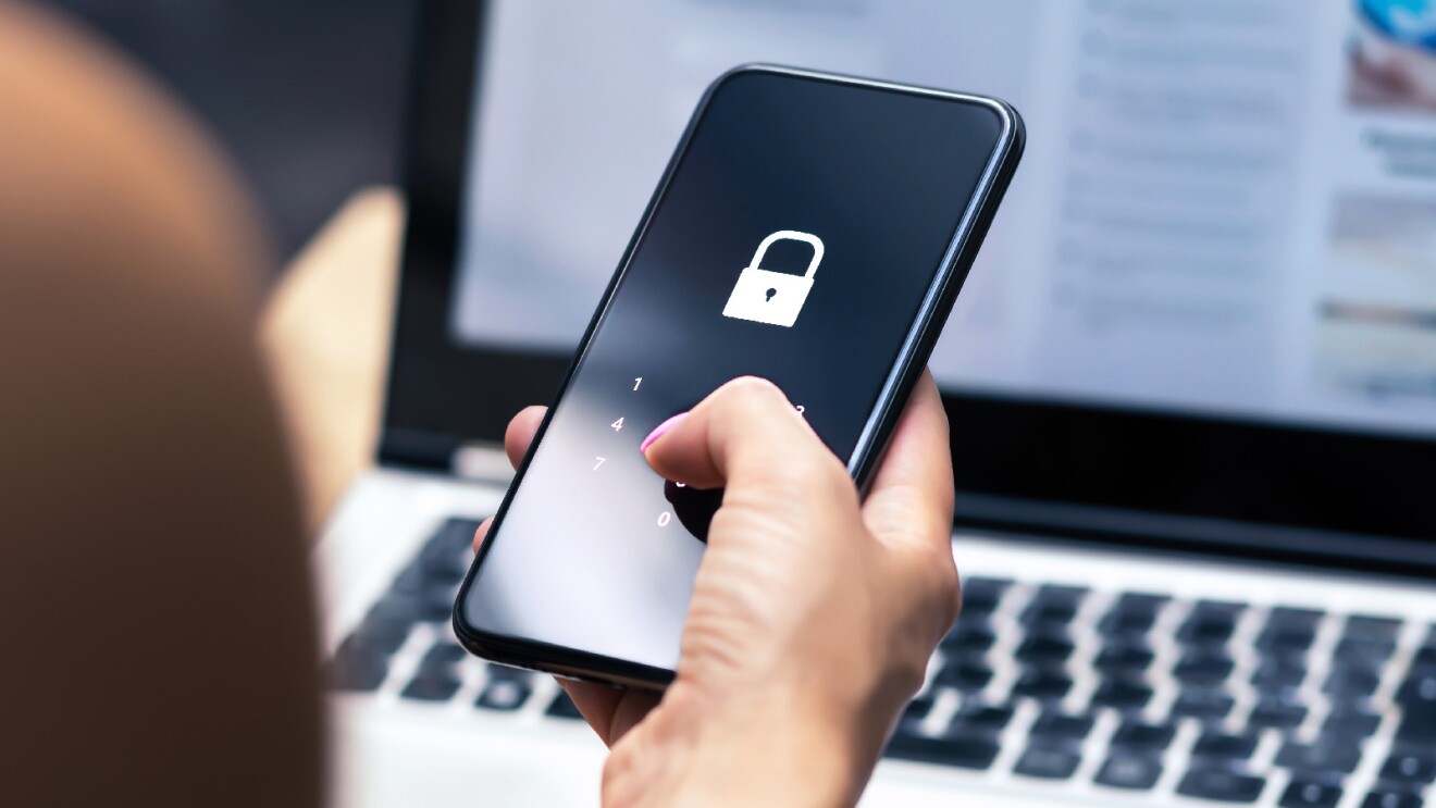 Prime Day 2023 live blog best deals: a woman's hand holding her phone with a locked screen and an open computer in the background.