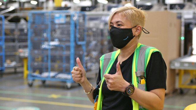 一名戴着口罩和安全背心的女子在镜头外对某人竖起了两个大拇指。她正站在亚马逊物流中心。