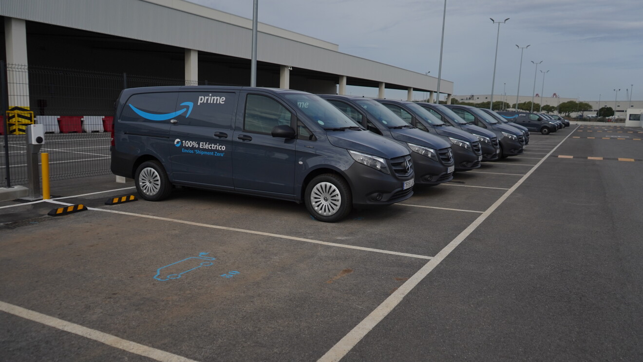 Una Mercedes Benz-Vans en un aparcamiento. Se ve el logo Prime de Amazon y el texto en la puerta 100% Eléctrico Envíos 'Shipment Zero'. En primer plano se ve un cargados de batería que está subministrando electricidad a la furgoneta de al lado que solo se ve un poco su parte trasera. 