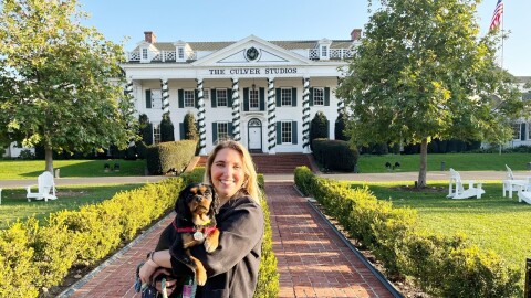 一个女人拿着一只狗的形象,微笑着站在面前的一个大大的白色殖民地风格建筑,说“斑鸠工作室”。