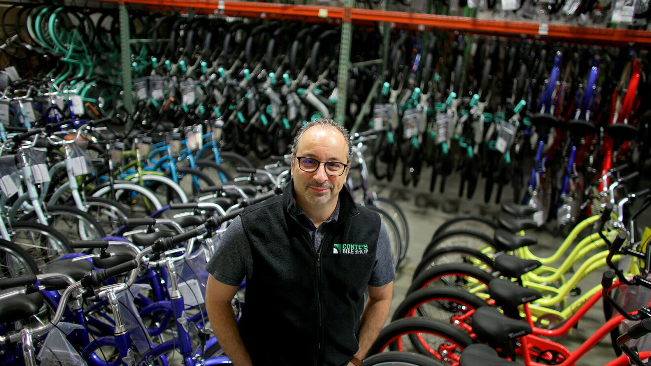 An image of a man smiling for a photo in a bike shop with rows of bikes behind him. He is wearing a vest that says "Conte's Bike Shop."