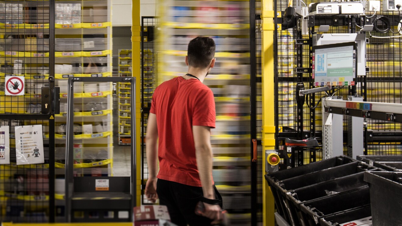Un dipendente Amazon ripreso di spalle al centro dell'immagine osserva gli scaffali mobili del magazzino.