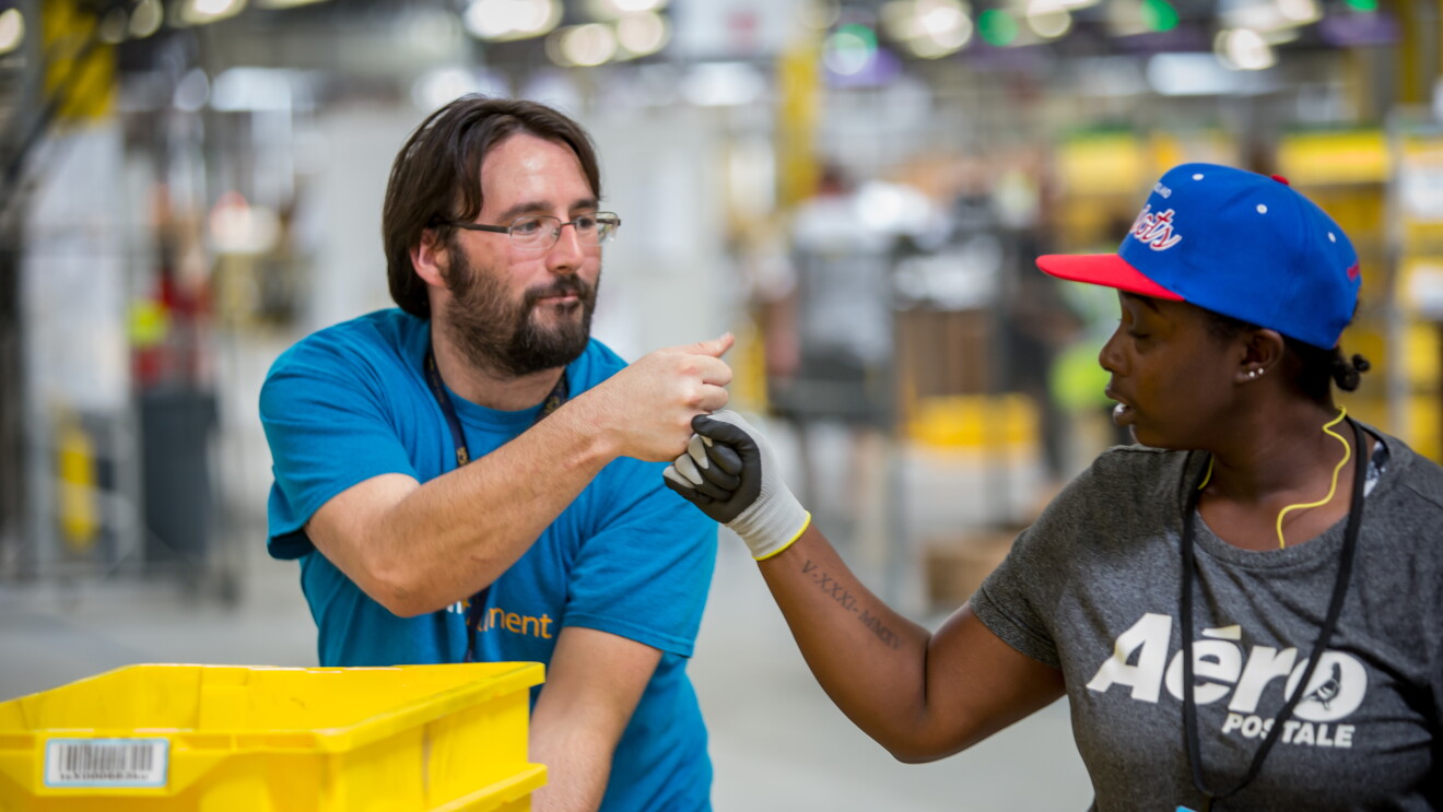 An male Amazon associate in a blue t-shirt bumps fists with a female Amazon associate wearing a grey t-shirt that says 'Aeropostale'. Three yellow bins are in front of the man, and the woman is wearing a blue baseball cap. 