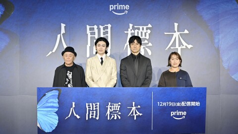 青い壁に「人間標本」の大きな文字。その前に3人の男性とその右側に女性1人が立っている。4人の前には、青い蝶の羽をあしらった青いパネルがあり、白い字で「人間標本」と書かれている。