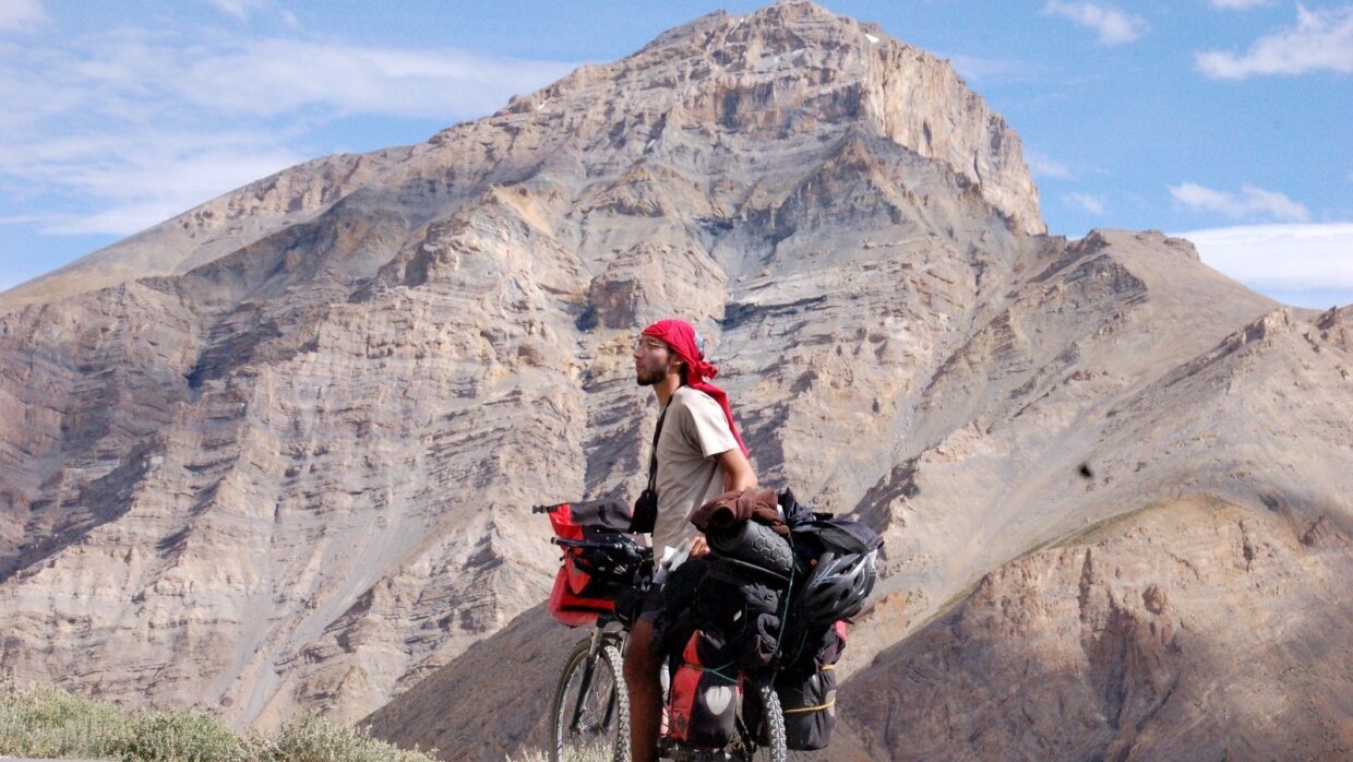 Karim Fahssis se tient à côté de son vélo chargé de sacs de voyage au milieu d'un paysage désertique. Il porte un foulard sur la tête. Derrière lui une montagne aride.