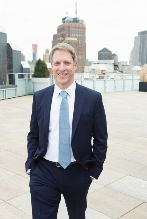 A man in a navy suit, white shirt, and slate blue tie stands outdoors in a city setting.