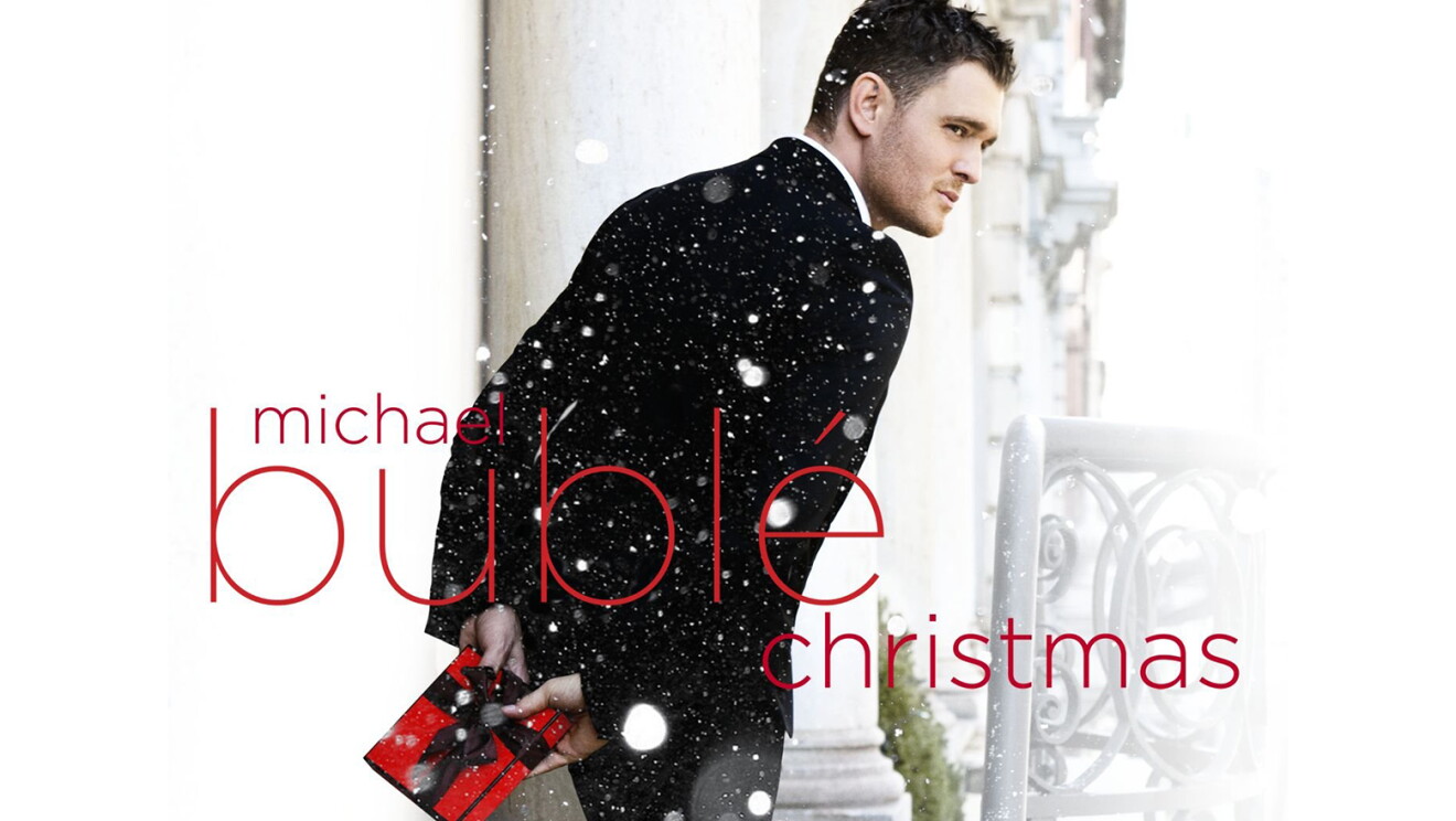 Michael Buble stands outside wearing a tuxedo, facing away, but seeming to be glancing over his shoulder. he holds a small, wrapped gift in red paper with black bow. Snow is falling in the image, and text saying "Michael Buble Christmas" is written over the top of the image.