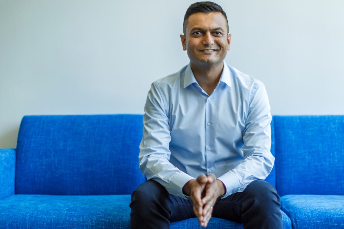 Amazon Music director of product management Kintan Brahmbhatt sits on a blue couch with his arms resting on his knees. He is wearing a blue shirt and jeans. 