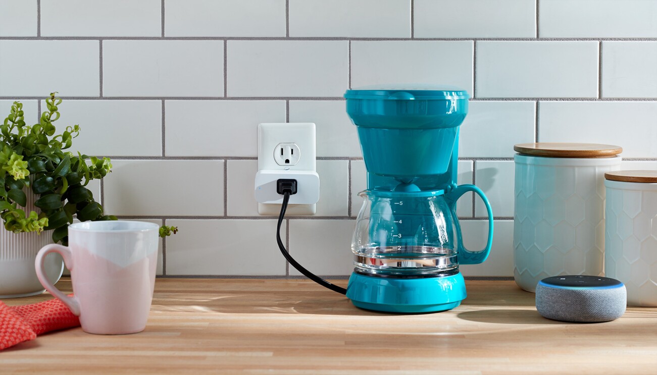 Amazon Smart Plug in a kitchen setting. It's plugged into an outlet, with a coffee pot plugged into it. To the right is a new Echo Dot device, and two stoneware storage containers. To the left is a potted plant, a coffefe pot and hand towel. 