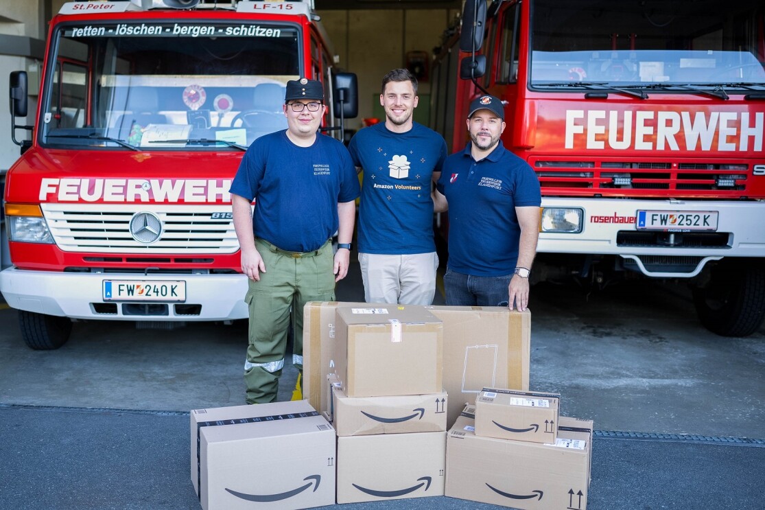 Amazon Mitarbeiter Andreas Widowitz mit Amazon Volunteers T-Shirt steht zusammen mit zwei Kollegen von der Freiwilligen Feuerwehr St. Peter mit Spenden in Amazon Kartons vor zwei Feuerwehrfahrzeugen.
