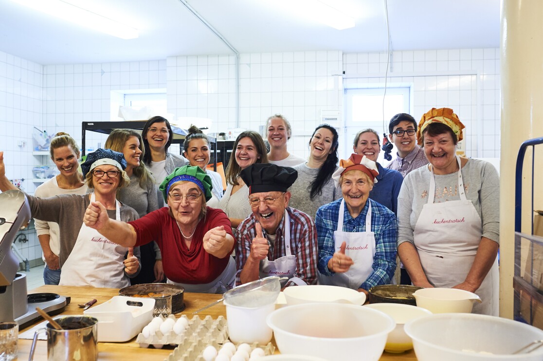 Das Team von Kuchentratsch steht in der Backstube und lacht in die Kamera. Sie tragen Schürzen und Kochmützen.
