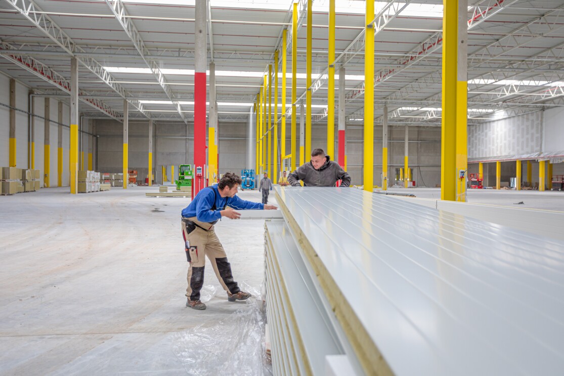 Eine leere Halle: im Vordergrund sind 2 Bauerbeiter beschäftigt, lange Dielen zu stapeln.