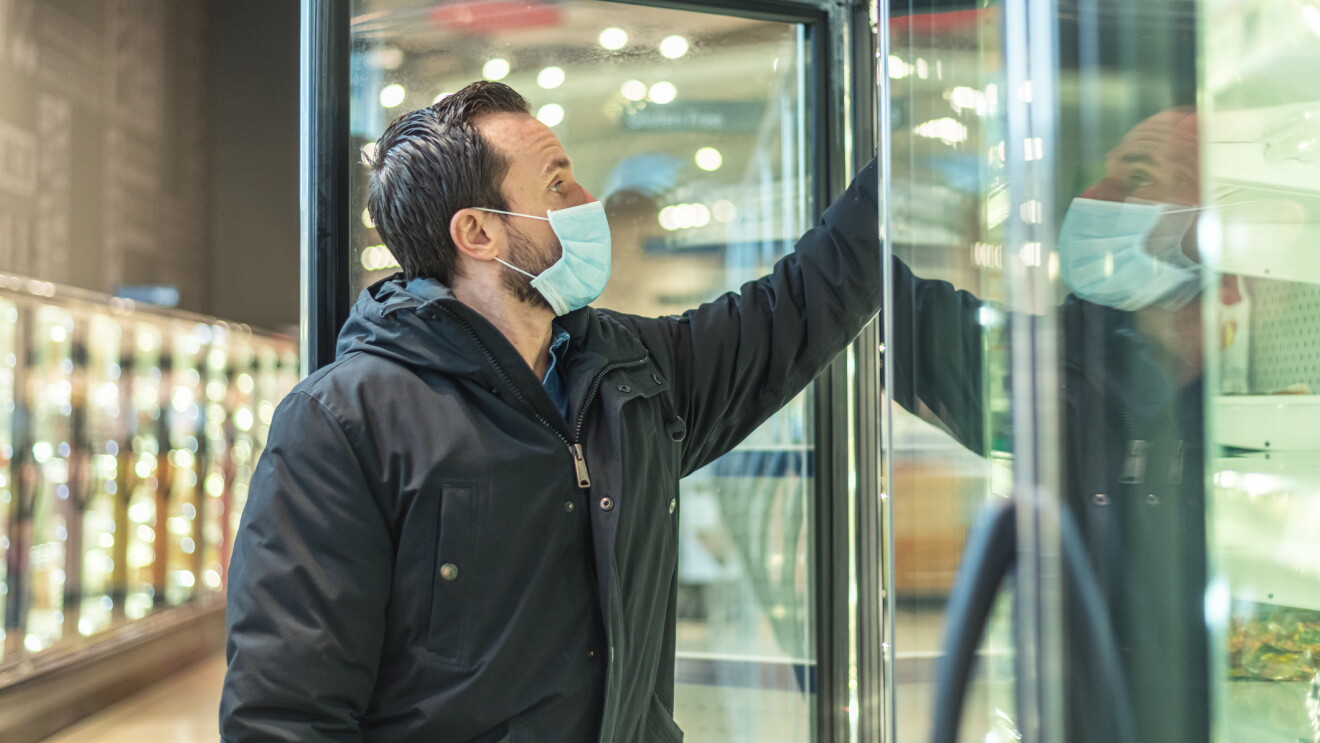 A man wearing a face mask stands at an open frozen foods cooler in a grocery store, reaching in to grab an item. 