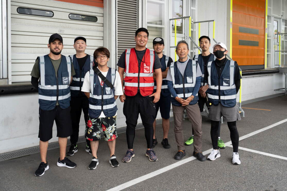 トラックが荷物を積み下ろしする場所で、赤いベストを着ている男性がネイビー色の安全ベストを着ている男女7人の中心に立っている