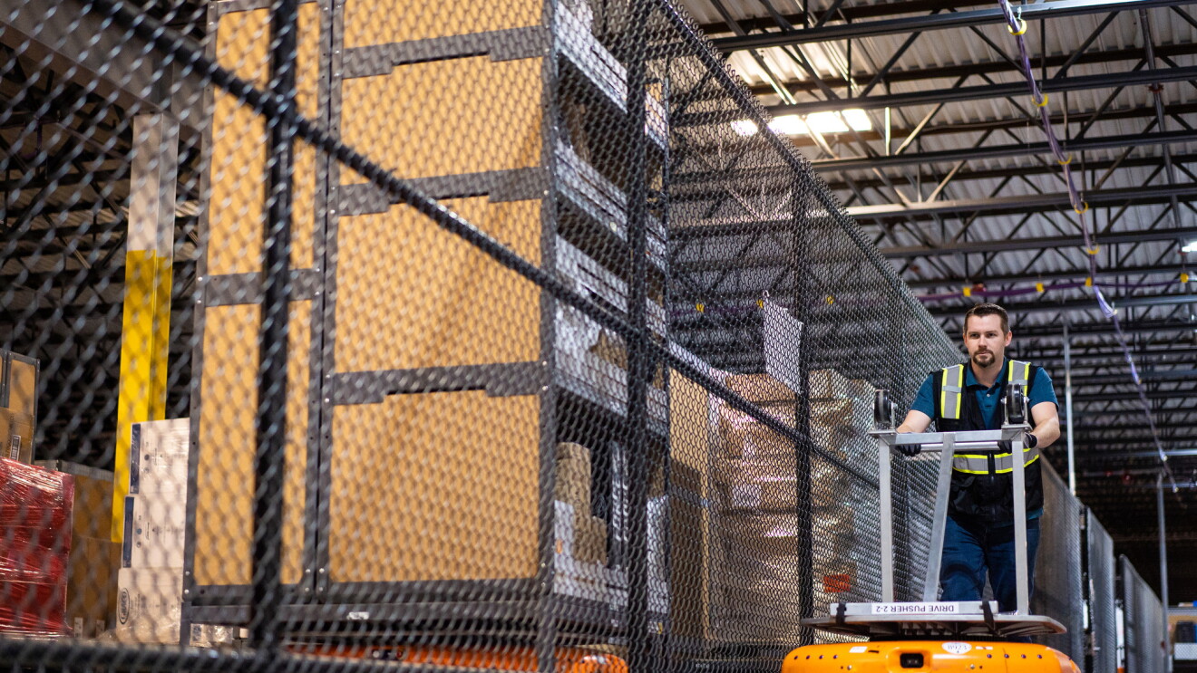 A man in a warehouse pushes a metal handle attached to a robot. The robot is orange and only about as high as the man's shins.