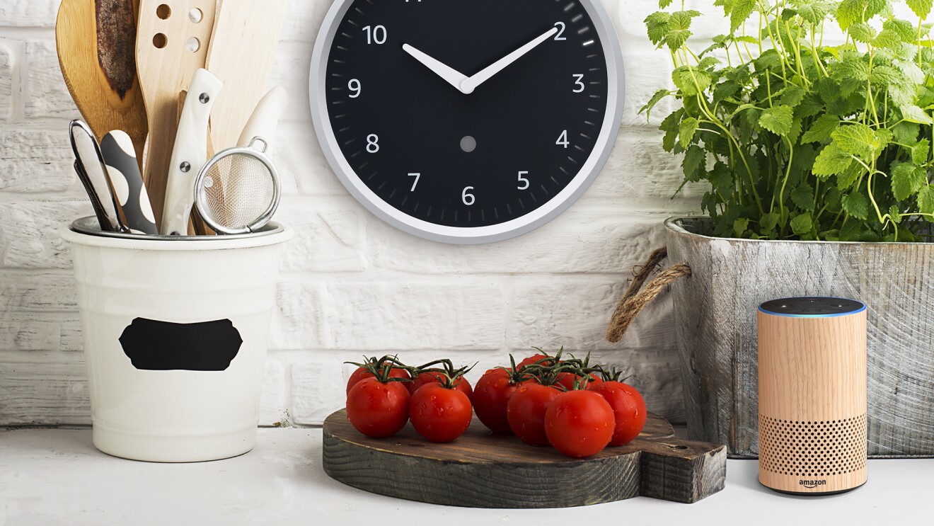 亚马逊回声在厨房墙上的钟。左边是厨房的玻璃水瓶工具,下面的时钟是一个砧板,西红柿。向右,一个草生长在一个容器,和一个回声设备。
