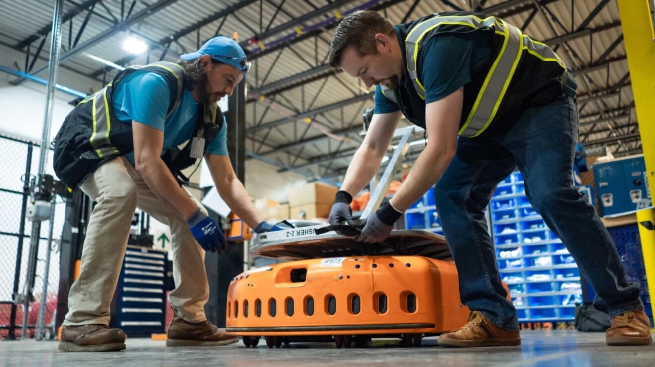 Two men in a warehouse space reach down to secure a silver-colored object to an orange, wheeled piece of machinery.