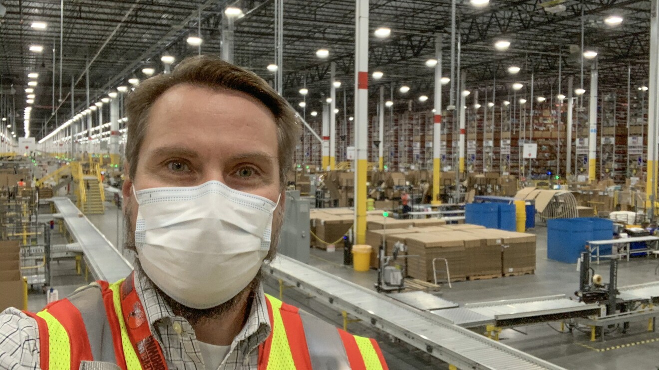 A man wearing a protective mask and a safety vest stands in front of a large warehouse space.