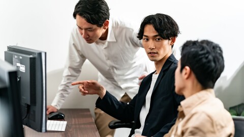 パソコンのモニターを前に3人の男性がカジュアルな感じで議論をしている