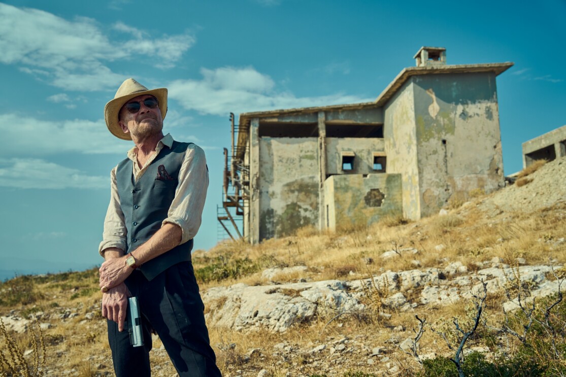 Man in cowboy hat and vest stands before dilapidated building in arid landscape