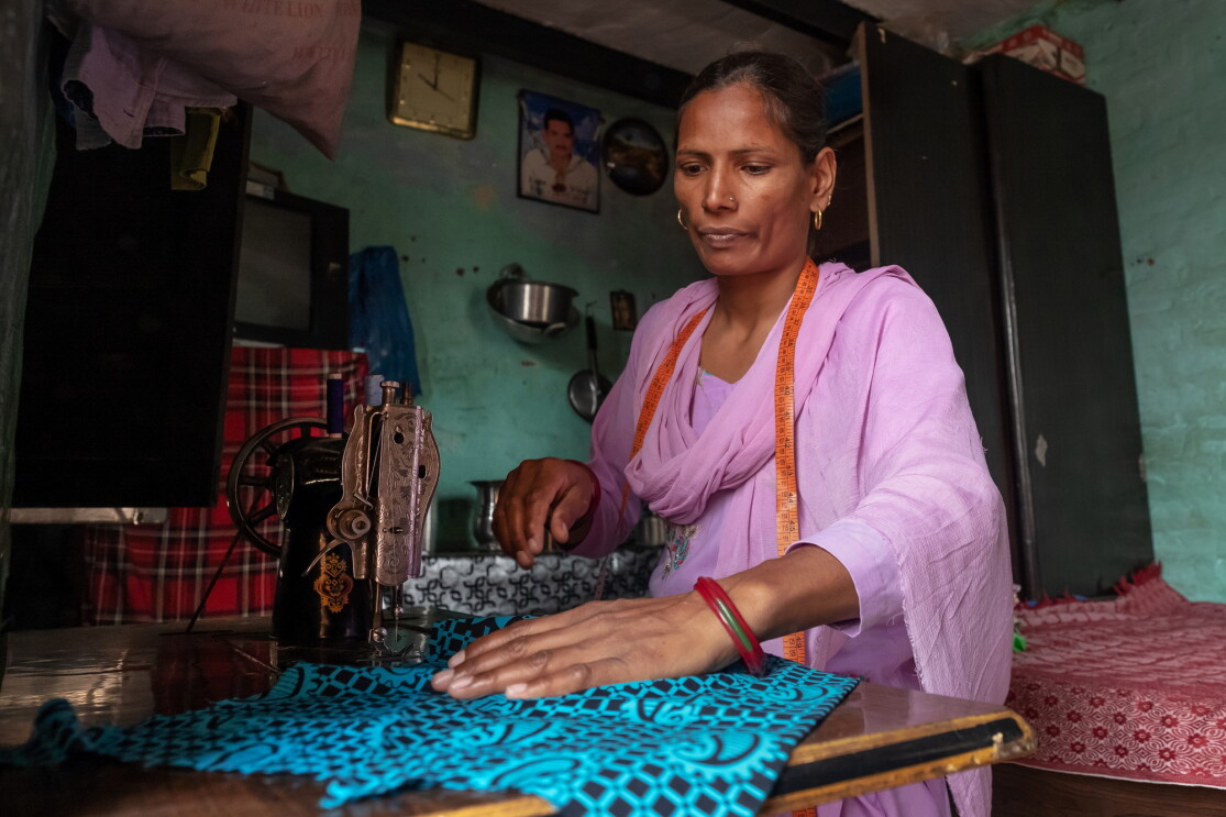 A woman sits at a table with a sewing machine. She has a measuring tape draped around her neck and uses her right hand to position a patterned fabric.