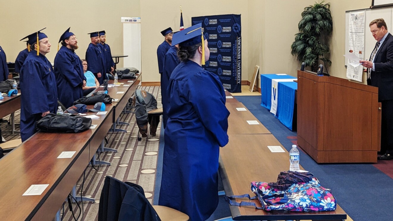 Graduates wear blue cap and gowns and stand while listening to a man talking at a podium.