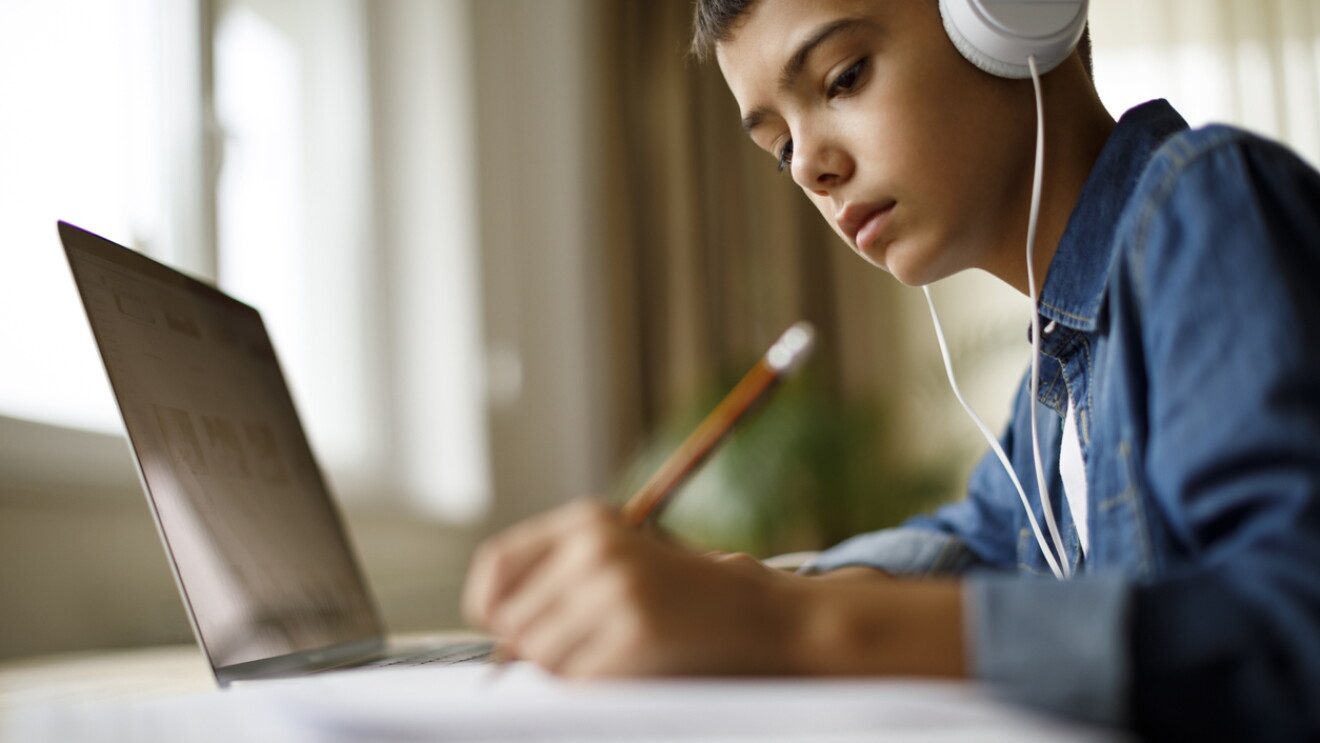 A student wearing headphones jots notes on a pad of paper as he sits in front of a laptop.