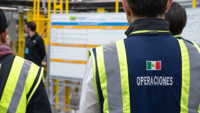 En el centro logístico Mex6 se ven difuminados y de espalda a tres trabajadores con sus chalecos de color amarillo y .verde. Detrás se ve el escudo de México. De fondo se ve a un hombre con el polo negro y dos pizarras escritas y con un plantilla azul y amarilla. 