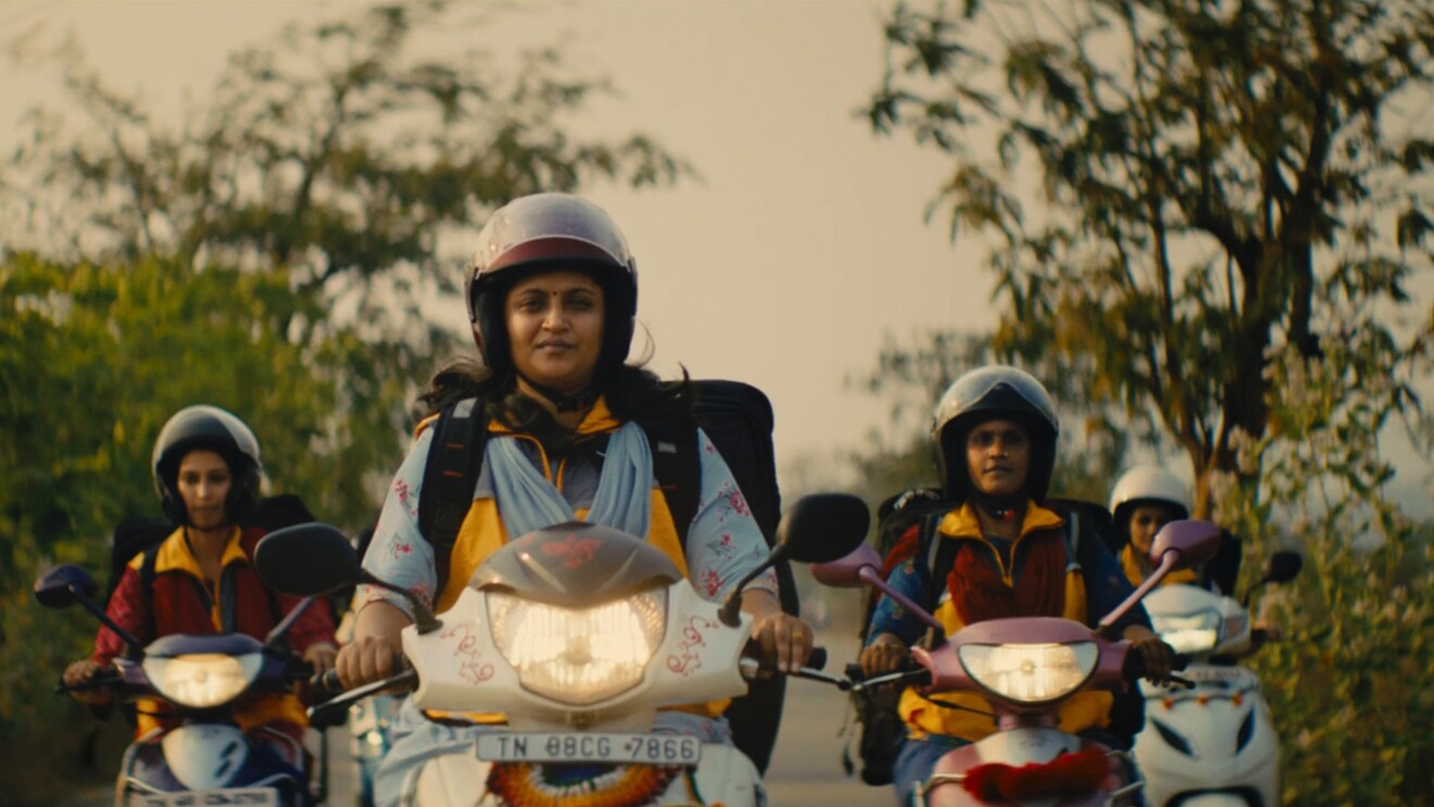 Cuatro mujeres indias en moto. Todas ellas llevan casco y una mochila grande donde cargan los pedidos. Las motos son dos de color blanco y dos de color morado y van con las luces endendidas. De fondo árboles.  y un cielo de color marrón. 