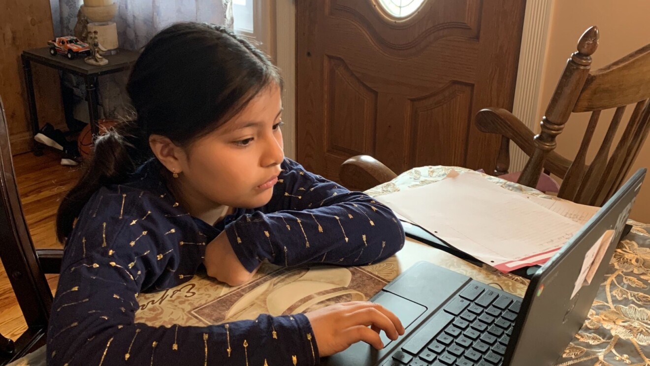 A girl sits a dining room table with a tablecloth and works on a laptop computer.