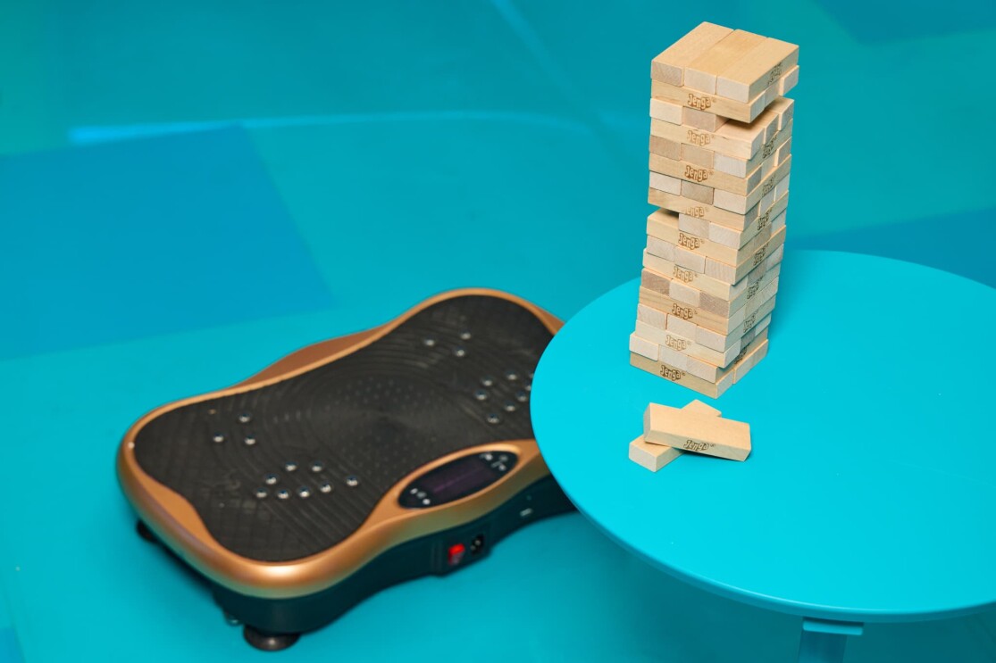 水色で統一された部屋。水色の丸テーブル上に木製のジェンガが積み上げられている。テーブルの下、水色の床にはブロンズ色の縁取りの黒い振動フィットネスマシンが置かれている。