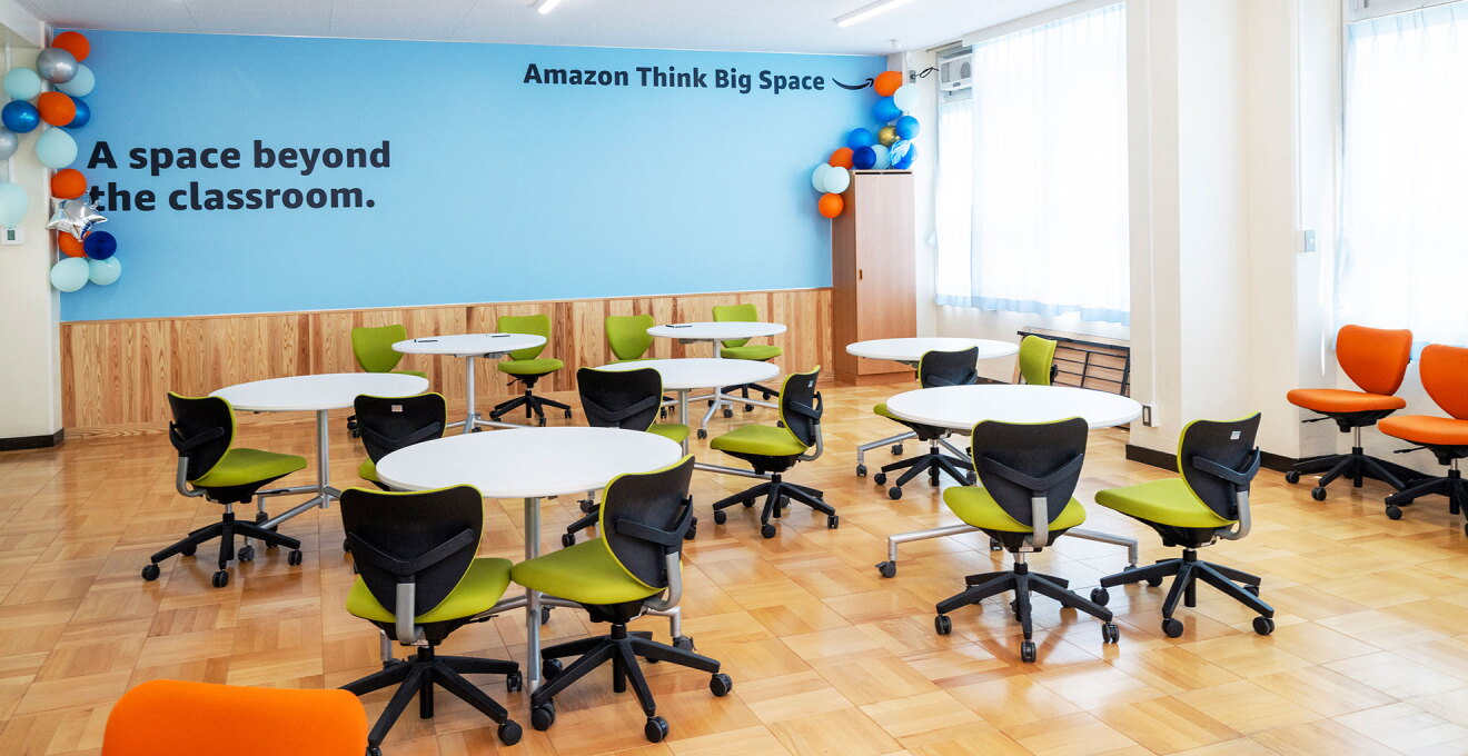 窓から自然光が入る明るい教室。水色の壁に「A space beyond the classroom.」、「Amazon Think Big Space」という黒文字。壁の両側天井付近にオレンジ、青、白の風船による装飾。白い丸テーブルが7つ、テーブルごとに背もたれと座面が黄緑色のキャスター付き椅子が2脚ずつ配置されている。