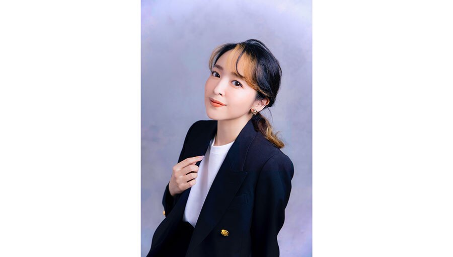 薄紫の背景に、濃紺のジャケットを着用した女性の上半身。部分的に茶髪の長髪を後ろでまとめている。