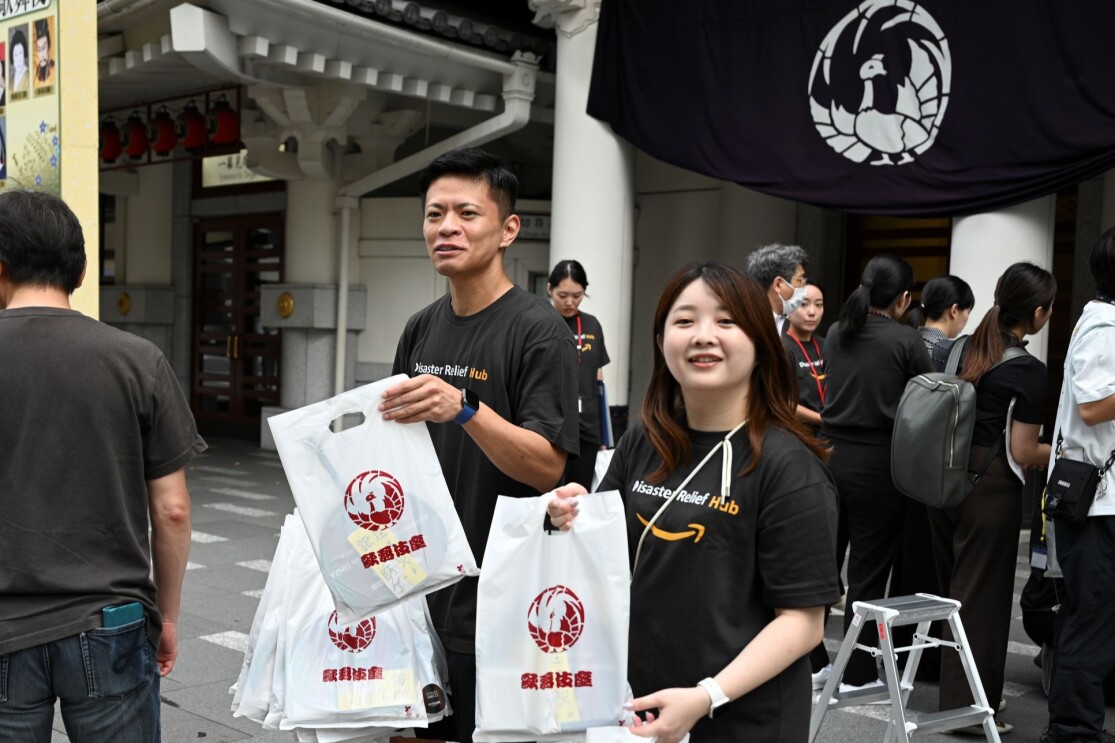 黒いAmazon Tシャツを着ている男性と女性が銀座歌舞伎座の前で、歌舞伎座のロゴを印刷した白い袋を配っている