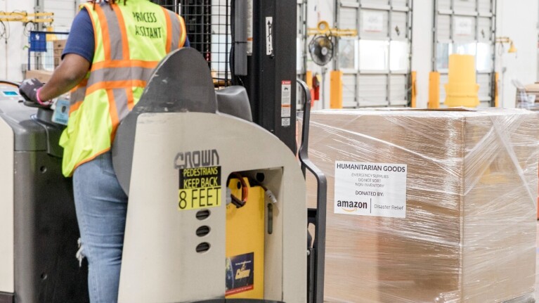 A woman wearing a yellow safety vest operates a fork lift to move a box that is labeled, 'humanitarian goods' at the disaster relief hub.