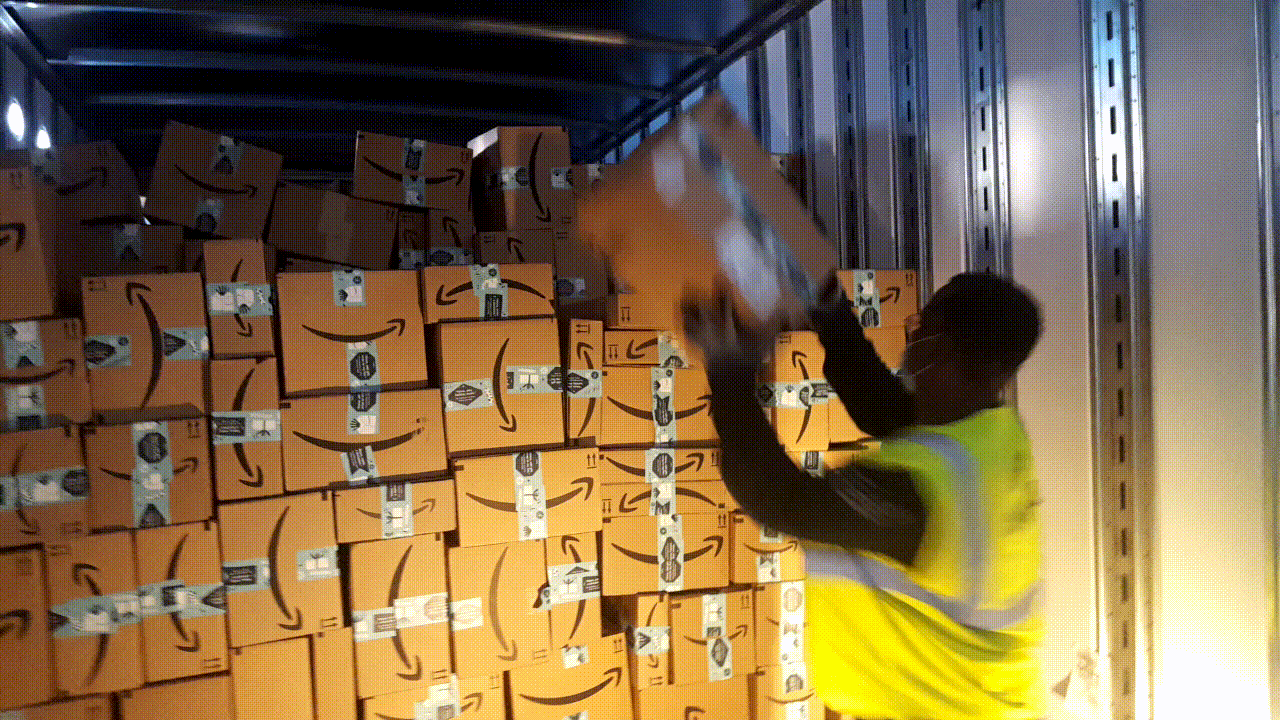 A Black man wearing a safety vest and mask loads a box into the back of an Amazon truck as he prepares it to move packages from an Amazon fulfillment center.