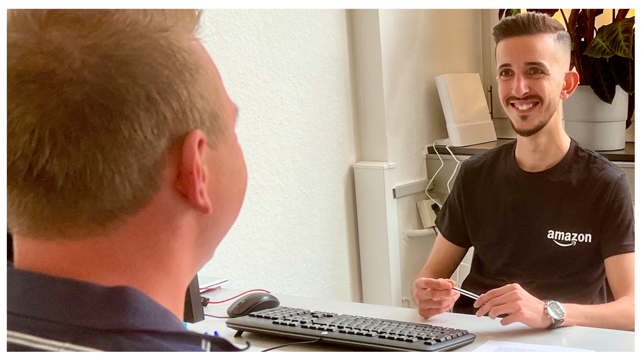Fuad, ein junger Mann in Amazon T-Shirt und mit braunen kurzen Haaren, sitzt an einem Schreibtisch und unterhält sich mit einem Bewerber. Vor ihm eine Tastatur und ein Rechner.